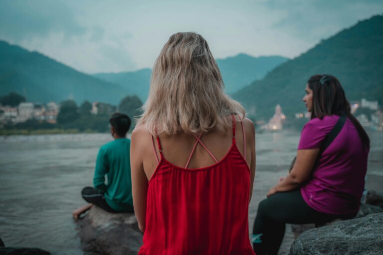 Three people meditate by the Ganges River in Rishikesh, India, with a serene mountain backdrop.