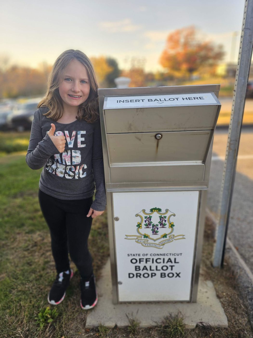 Adeline giving a thumbs up beside a Connecticut official ballot drop box