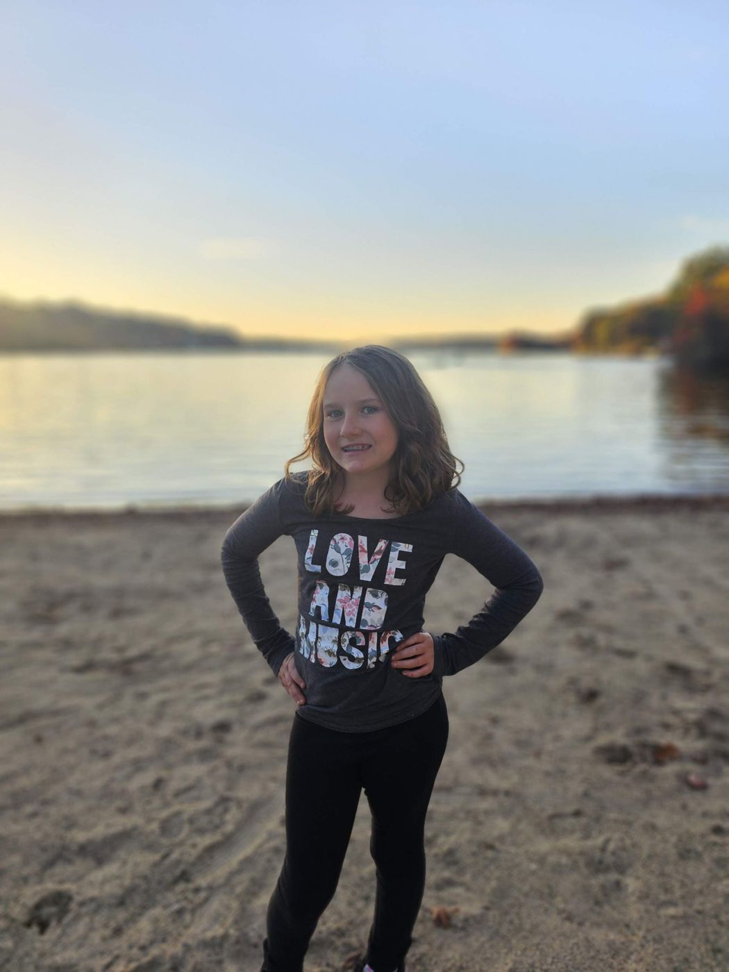 Adeline smiling on a lakeside beach at sunset in a 'Love and Music' shirt